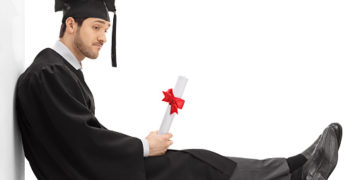 Sad graduate student with a diploma sitting on the floor and leaning on a wall isolated on white background